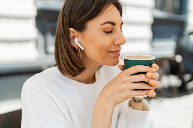 Blissful short haired woman enjoing cappucino in cafe, wearing cozy white sweater and listening to favorite music by earphones