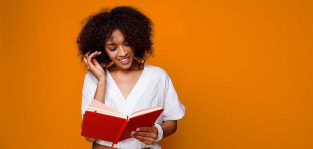 Blissful   smiling  mix race woman  reading with pleasure book  over orange background. Copy space for text.