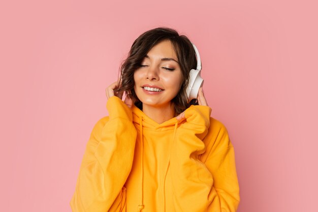 Blissful smiling woman in white earphones listening to music on pink. Wearing orange hoodie.