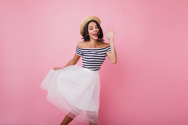 Blissful tanned woman waving hand while dancing. Indoor portrait of pleased brunette girl wears elegant lush skirt.