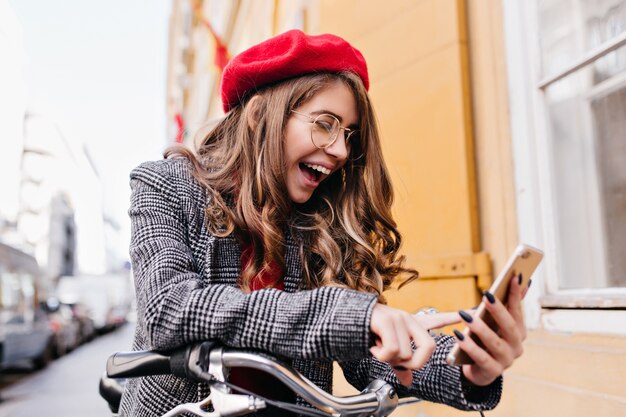 Blissful white woman with brunette hair looking at phone screen with smile on street background