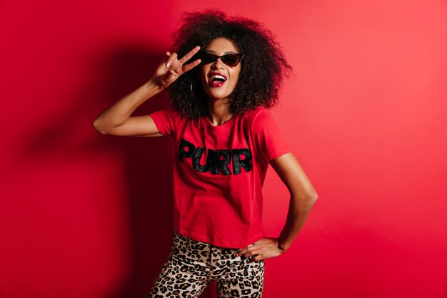 Blissful woman in trendy sunglasses laughing on red wall