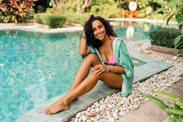 Blissful woman  with wavy hairs in  summer outfit sitting near pool  Vacation and travel mood Bright colors Trendy accesorises