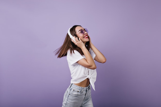 カジュアルな白いTシャツのダンスで至福の若い女性。ヘッドフォンでリラックスした流行の服を着た恍惚とした白人の女の子。