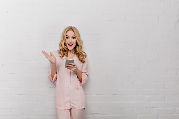 Blissful young woman in cotton sleepwear posing with phone in hands. Indoor photo of charming blonde female model in pink pajamas isolated on white wall.