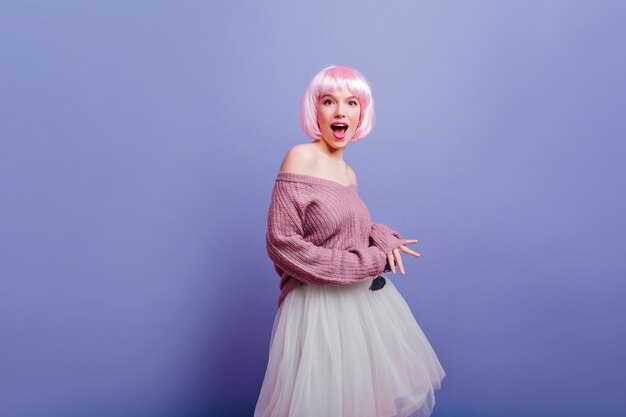 Blissful young woman wears white skirt and pink periwig posing with happy face expresshion. Pretty female model in fashionable peruke having fun 