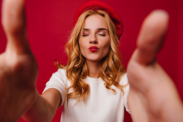 Blithesome beautiful girl in beret posing with kissing face expression. Indoor shot of adorable pretty woman in french outfit standing on red wall.