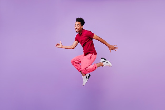 Blithesome black brunette man dancing with happy smile. Indoor photo of inspired guy in red pants and white shoes jumping.