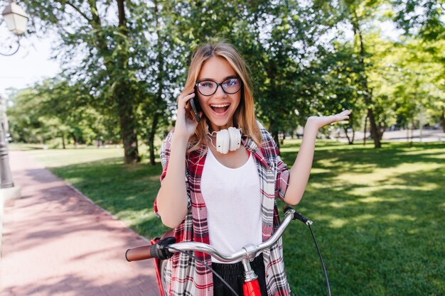 Blithesome blonde girl laughing during bike ride. Outdoor photo of ecstatic white woman in headphones.