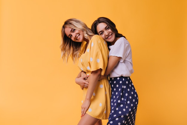 Веселая брюнетка женщина обнимает своего лучшего друга. Очаровательные сестры в летней одежде позируют на ярко-желтом.