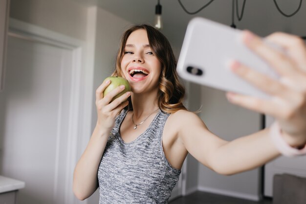 りんごで自分の写真を撮る陽気な白人の女の子。自撮りにスマートフォンを使って笑う愛らしい女性。