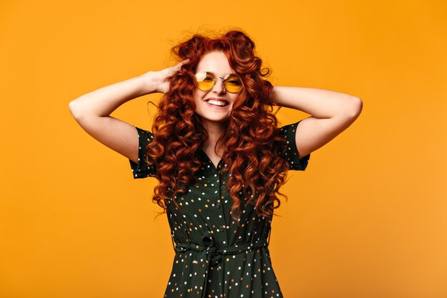 Blithesome european girl touching ginger hair and laughing. Studio shot of cheerful young woman in sunglasses posing on yellow background.