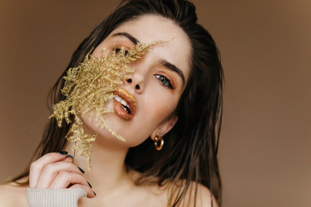 Blithesome european woman posing on brown wall with plant. Close-up shot of carefree girl with green leaf.