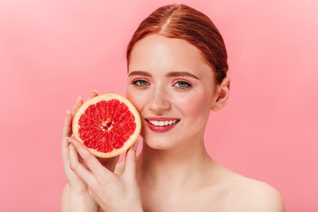 Blithesome ginger girl holding juicy grapefruit. Pretty young woman expressing happiness on pink background.
