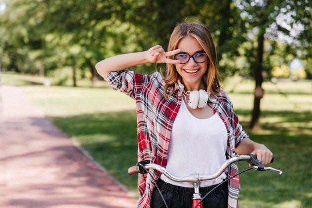 公園の周りに乗っている大きなヘッドフォンで明るい女の子。自然の中で自転車に座って笑っている愛らしい女性の屋外写真。