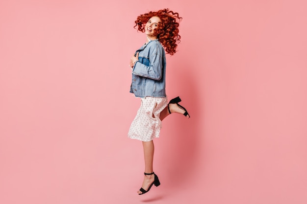 Free photo blithesome girl in dress dancing on pink background. full length view of excited ginger woman in denim jacket jumping with smile.
