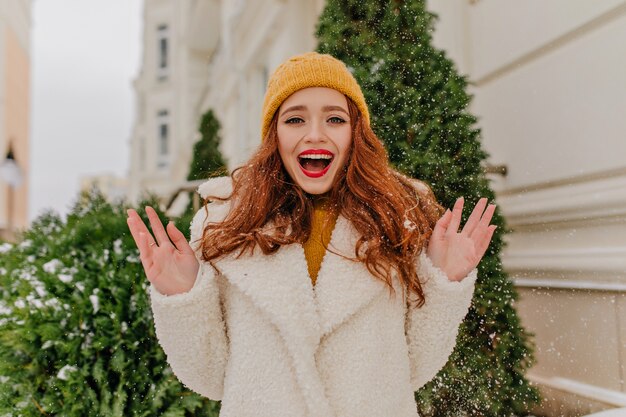 Blithesome long-haired woman enjoying winter weekend. Outdoor photo of excited ginger girl fooling around on the street.