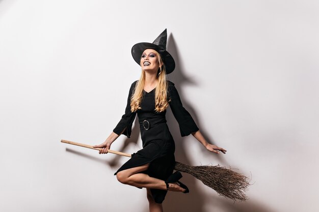 Blithesome long-haired woman having fun in halloween. Cute blonde girl in witch hat smiling at carnival party.