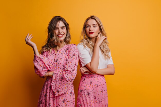 Blithesome white girl in dress relaxing with best friend. Indoor shot of glad caucasian sisters isolated on yellow wall.