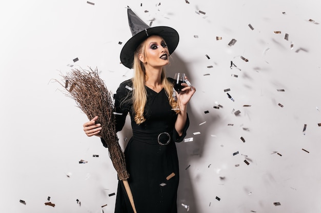 Blithesome young witch drinking wine. Carefree woman in halloween attire posing at party.