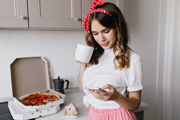 キッチンでコーヒーとテキストメッセージを飲む陽気な若い女性。朝食時に自宅でポーズをとる恍惚とした巻き毛の女性モデル。