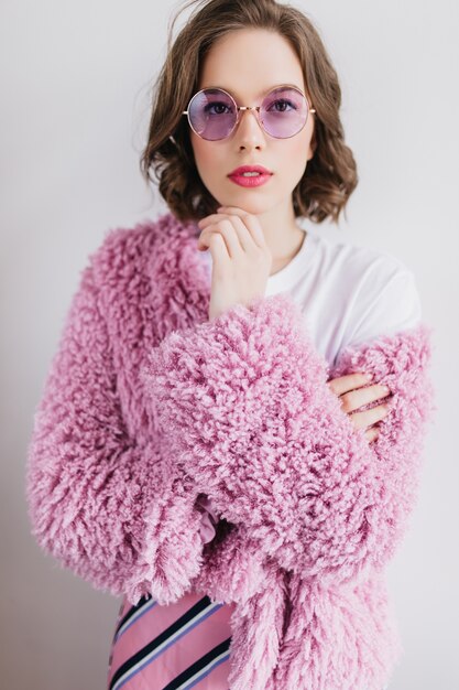 白い壁に立っているトレンディな丸いメガネの明るい若い女性。毛皮のコートを着たjocundの短い髪の女性モデル。
