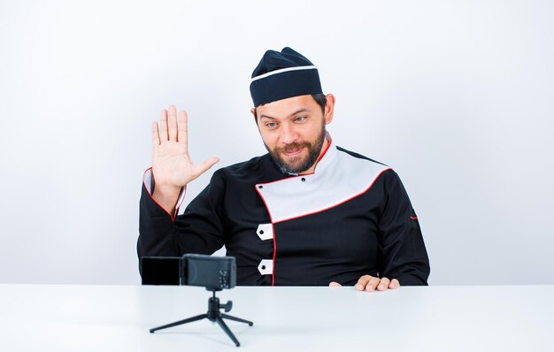 Шеф-повар блогера показывает приветственный жест, сидя перед своей мини-камерой на белом фоне