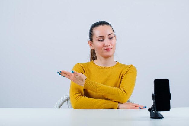 Blogger girl is pointing left with hand by sitting in front of mobile camera on white background