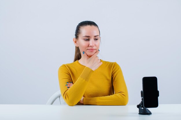 Blogger girl is putting hand on chin by sitting in front of mobile camera on white background