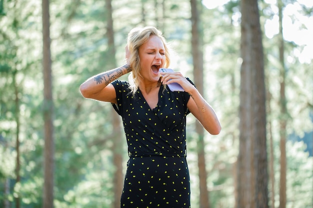 Blogger girl is signing by holding mobile as microphone on nature background