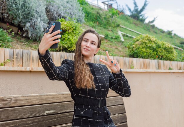 Девушка-блогер делает селфи со своим смартфоном, держа жест победы в парке