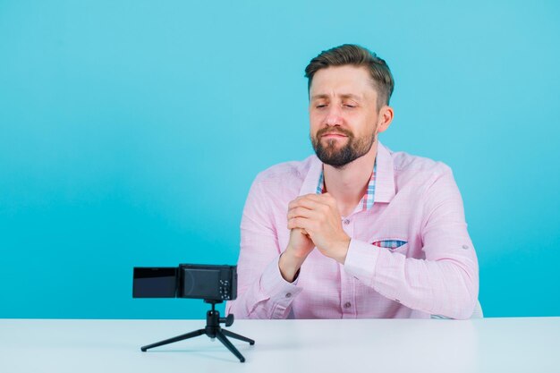 Blogger man is holding hands near chest by sitting in front of his mini camera on blue background