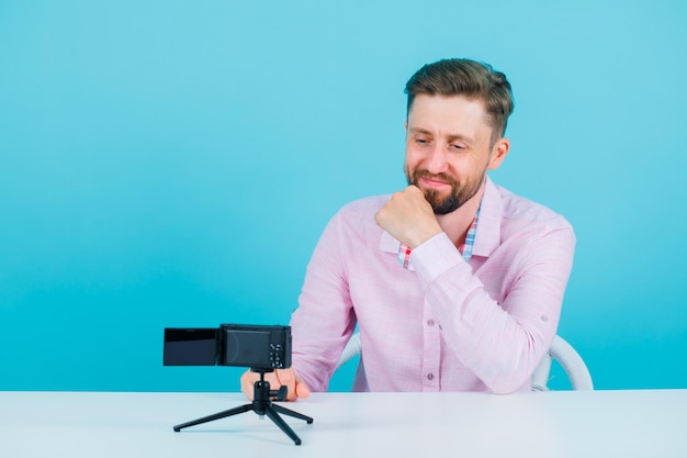 Blogger man is looking at his mini camera by holding hand on chin on blue background