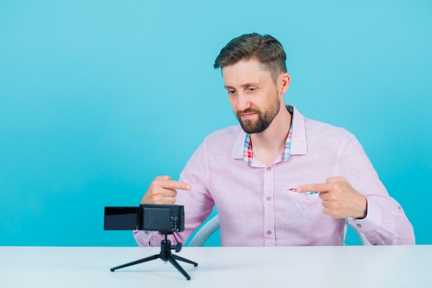 Blogger man is posing to his mini camera by pointing himself with forefingers on blue background