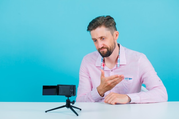 Blogger man is posing to his mini camera by pointing left with hand on blue background