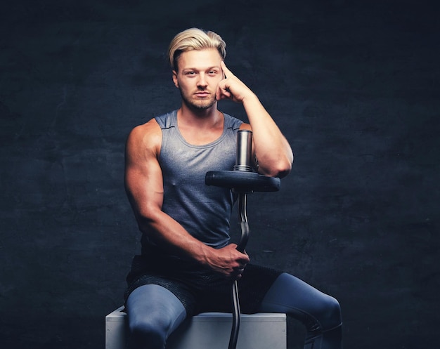 Free photo a blond, athletic male dressed in a grey sportswear sits on a white wooden box and holds barbel.