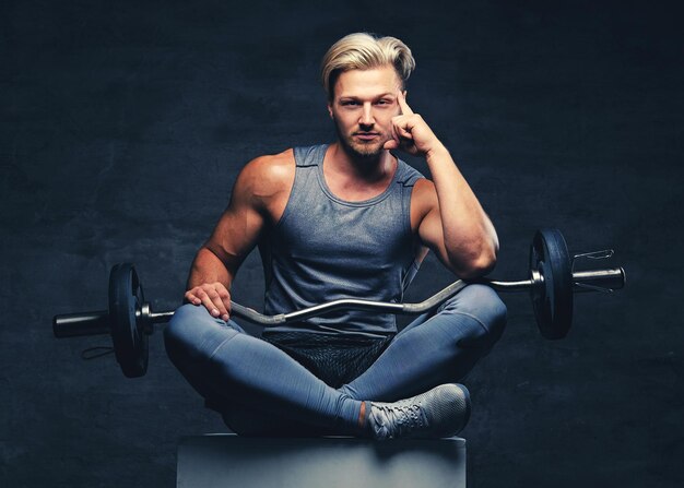 A blond, athletic male dressed in a grey sportswear sits on a white wooden box and holds barbel.