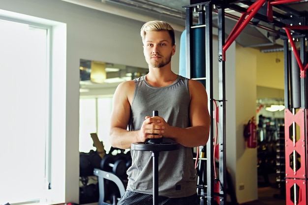 Blond athletic male in sportswear holds barbell over TRX stand background in a gym club.
