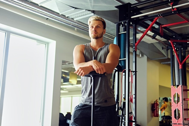 Блондин спортивный мужчина в спортивной одежде держит штангу на фоне стенда TRX в спортзале.