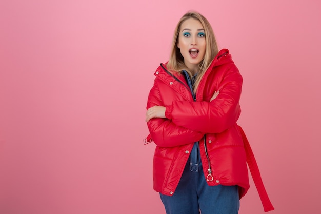 Blond attractive active woman posing on pink wall in colorful winter down jacket of red color, having fun, warm coat fashion trend, surprised shocked face expression