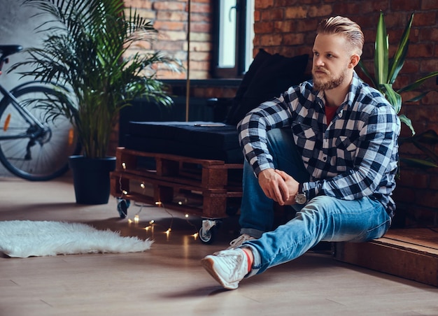 A blond bearded hipster male dressed in a jeans and fleece shirt posing in a room with loft interior.