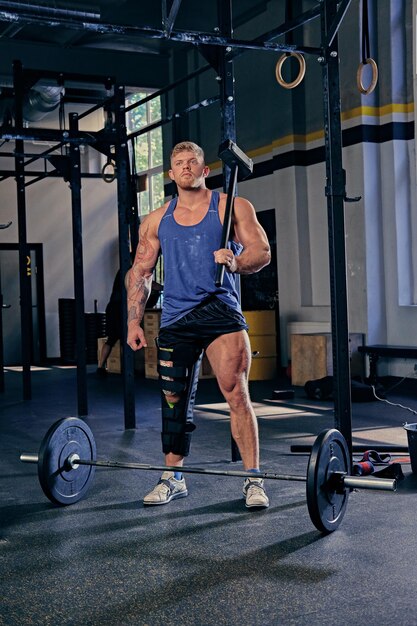 Blond bodybuilder with broken leg in bandage holds cross fit hammer in a gym club.