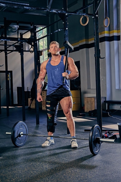 Blond bodybuilder with broken leg in bandage holds cross fit hammer in a gym club.