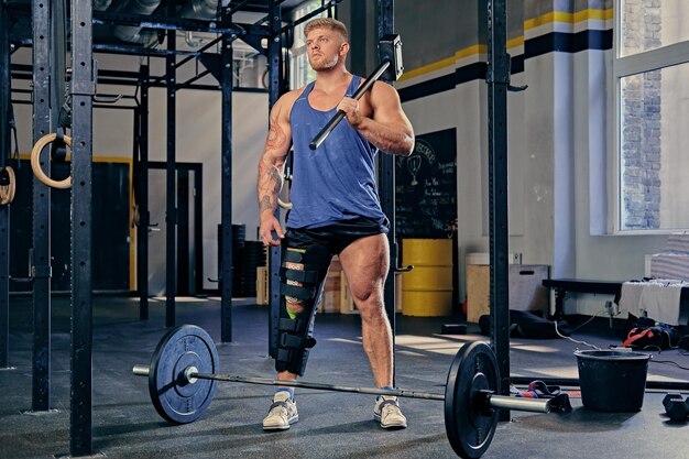 Blond bodybuilder with broken leg in bandage holds cross fit hammer in a gym club.