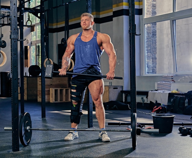 Blond bodybuilder with broken leg in bandage holds cross fit hammer in a gym club.