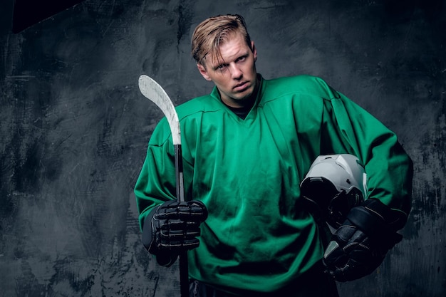 Free photo the blond caucasian hockey player holds protective helmet and playing stick on grey background.