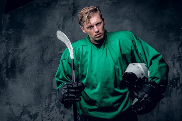 Foto gratuita il giocatore di hockey biondo caucasico tiene un casco protettivo e un bastone da gioco su sfondo grigio.