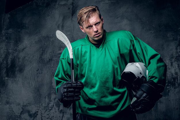 The blond Caucasian hockey player holds protective helmet and playing stick on grey background.