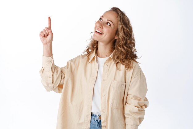 Blond cute girl pointing and looking up at promotional text, showing banner, standing over white wall