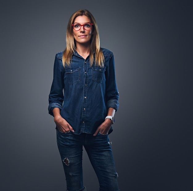 Blond female dressed in denim shirt and red eyeglasses posing over grey background.